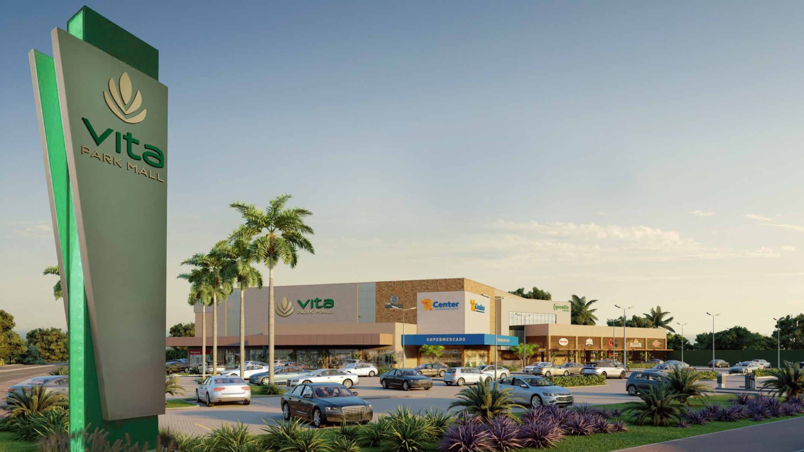 Totem verde do Vita Park Mall na entrada, com palmeiras e o estacionamento em frente ao shopping